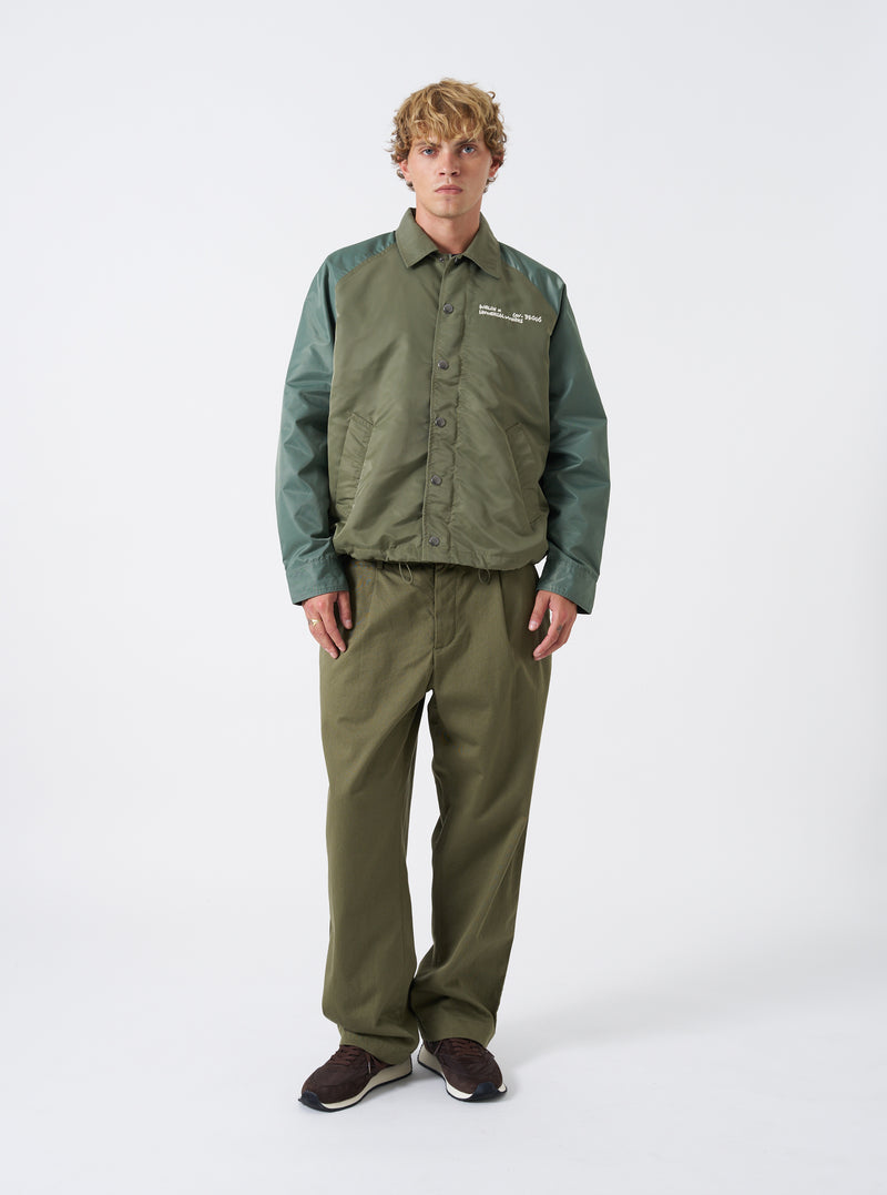 A male model wears the Walsh x Universal Works Olive Flight Nylon Club Coach Jacket against a neutral studio backdrop. The shot highlights the relaxed fit, front snap fastenings, classic shirt collar, and olive green flight nylon fabric.