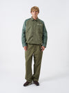 A male model wears the Walsh x Universal Works Olive Flight Nylon Club Coach Jacket against a neutral studio backdrop. The shot highlights the relaxed fit, front snap fastenings, classic shirt collar, and olive green flight nylon fabric.