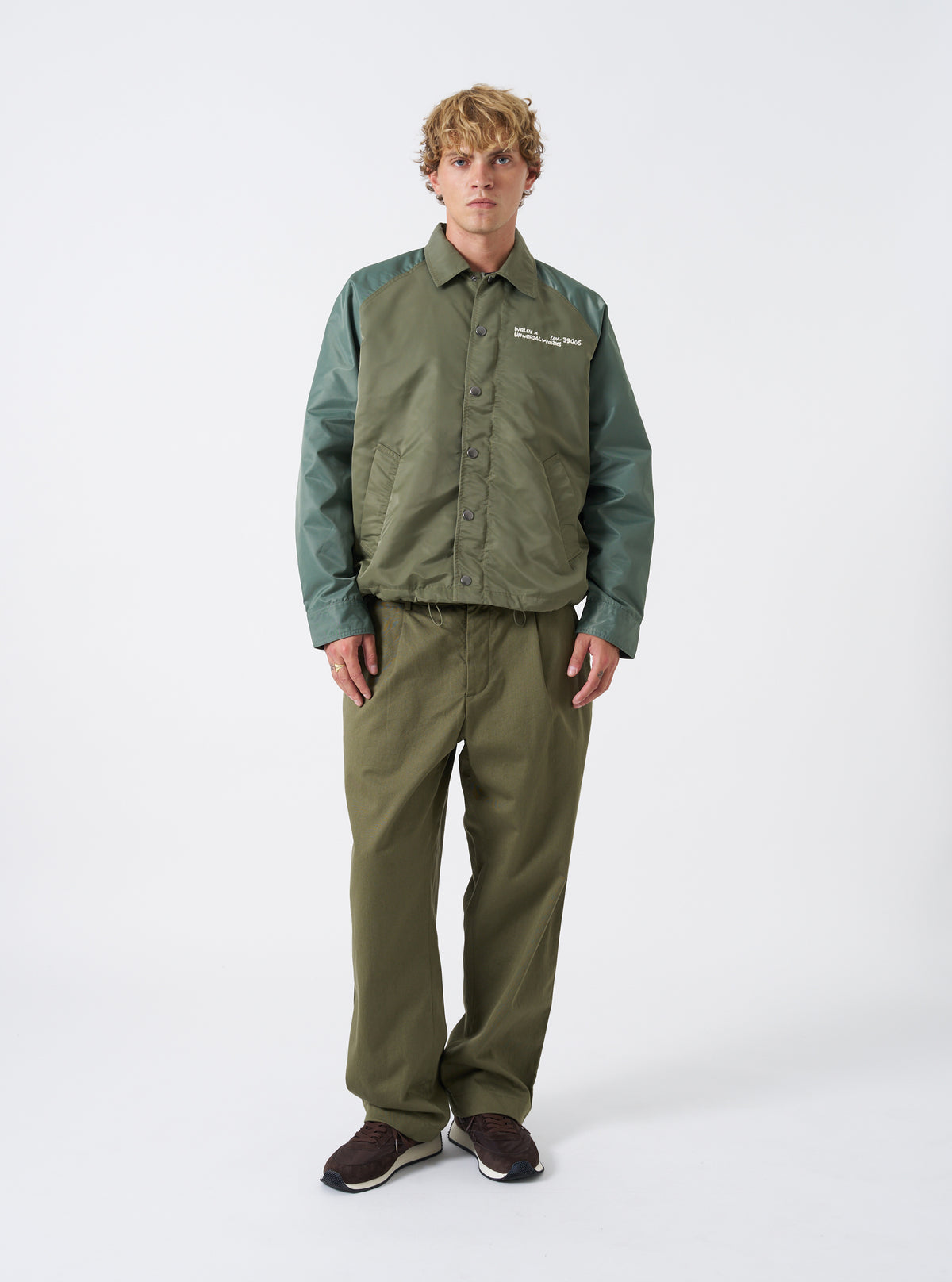 A male model wears the Walsh x Universal Works Olive Flight Nylon Club Coach Jacket against a neutral studio backdrop. The shot highlights the relaxed fit, front snap fastenings, classic shirt collar, and olive green flight nylon fabric.