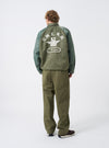 Full-length view of a model wearing the Walsh x Universal Works Olive Flight Nylon Club Coach Jacket styled with casual trousers in a studio. The image displays the comfortable fit, olive tone, side pockets, and snap-front closure.