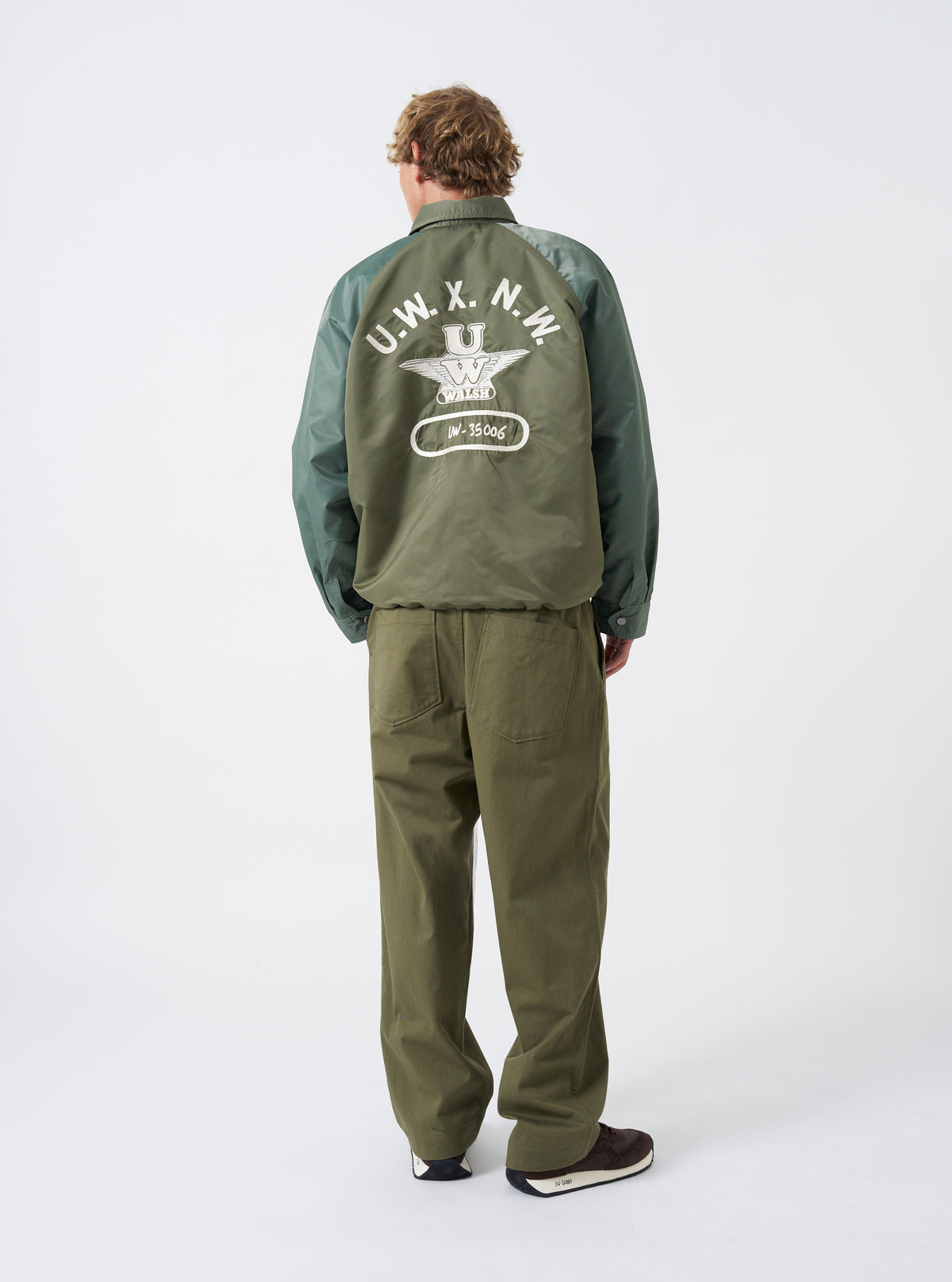 Full-length view of a model wearing the Walsh x Universal Works Olive Flight Nylon Club Coach Jacket styled with casual trousers in a studio. The image displays the comfortable fit, olive tone, side pockets, and snap-front closure.