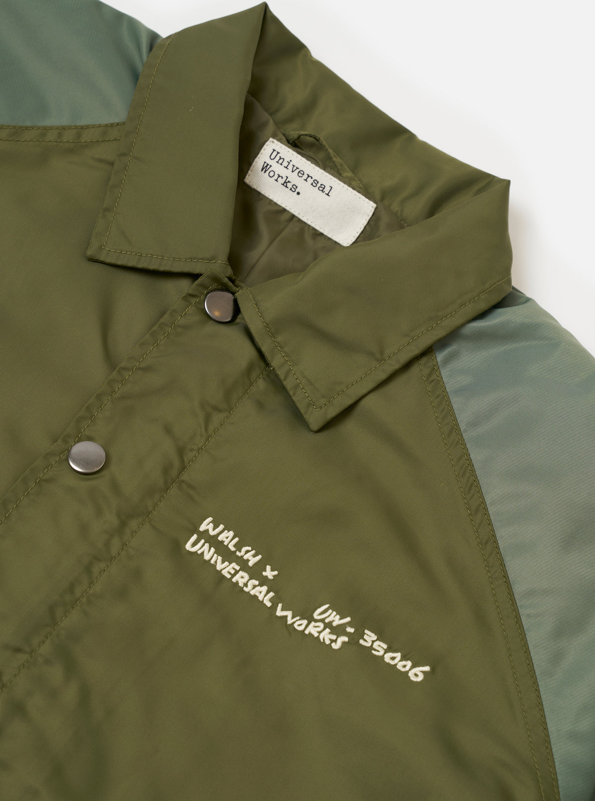 Front view of the Walsh x Universal Works Olive Flight Nylon Club Coach Jacket presented on a plain white studio background. The jacket features classic snap buttons, side welt pockets, a flat collar, and smooth olive green nylon fabric.