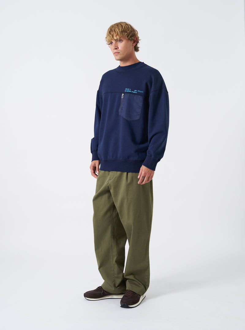 A model wearing the Walsh x Universal Works Navy Brushback Sweat Panelled Loose Sweatshirt stands against a plain studio backdrop. Prominent details include the relaxed loose silhouette, ribbed crew neck, ribbed cuffs, and navy tone.