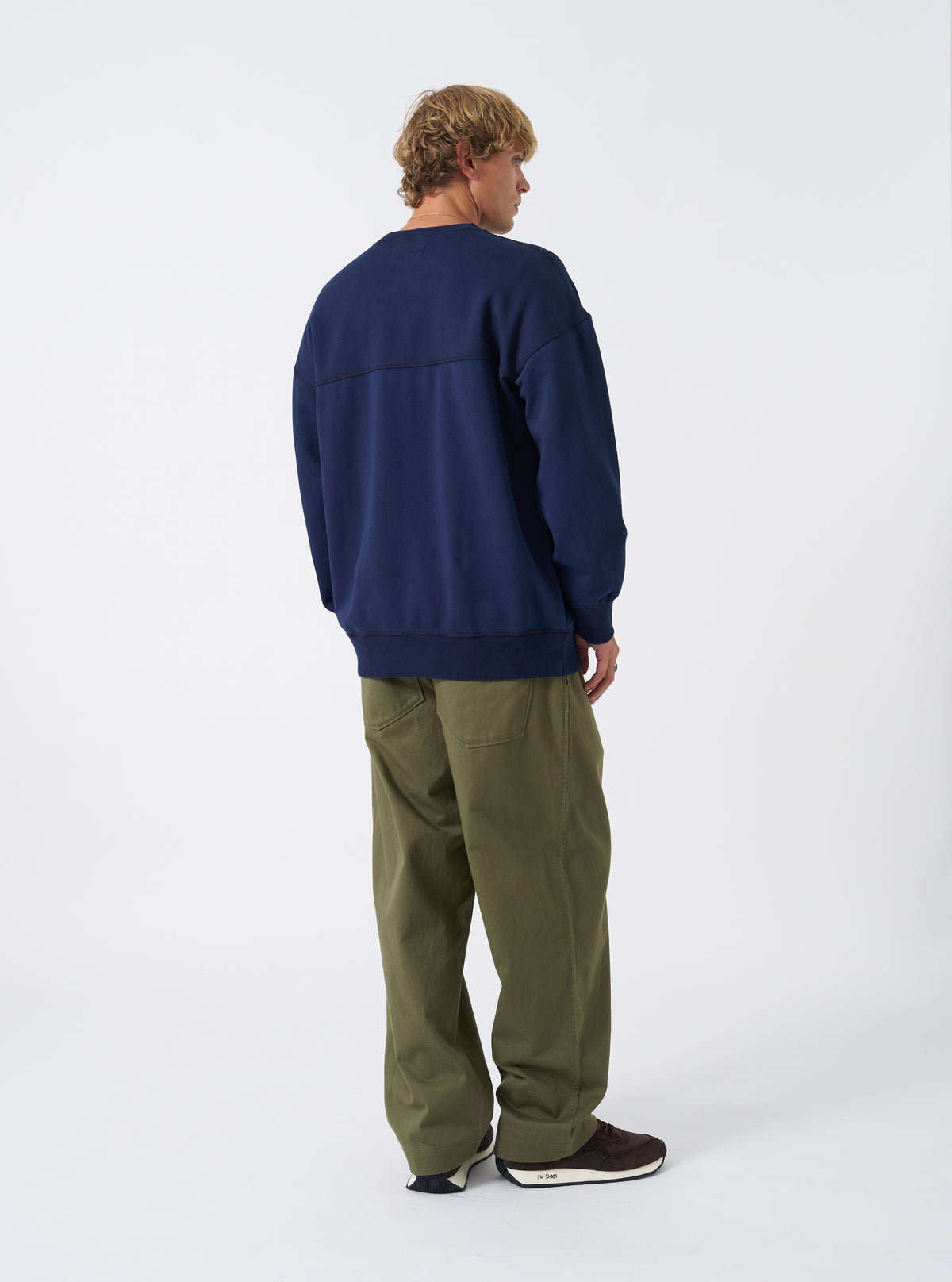 Side profile view of a model wearing the Walsh x Universal Works Navy Brushback Sweat Panelled Loose Sweatshirt in a studio setting. The image showcases the paneled construction, comfortable oversized fit, and soft brushback cotton fabric.