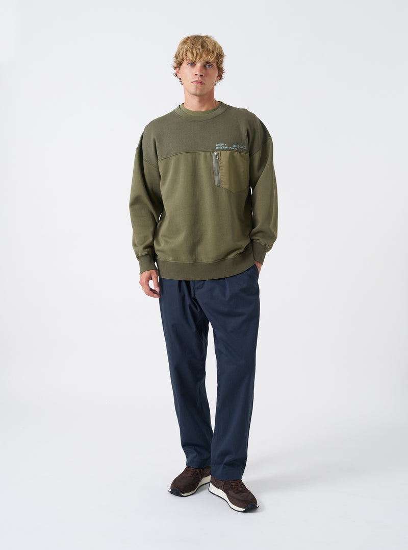 A model wears the Walsh x Universal Works Olive Brushback Sweat Panelled Loose Sweatshirt against a plain studio background. Prominent details are the relaxed fit, olive green tone, crew neckline, and soft brushback cotton texture.