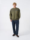 A model wears the Walsh x Universal Works Olive Brushback Sweat Panelled Loose Sweatshirt against a plain studio background. Prominent details are the relaxed fit, olive green tone, crew neckline, and soft brushback cotton texture.