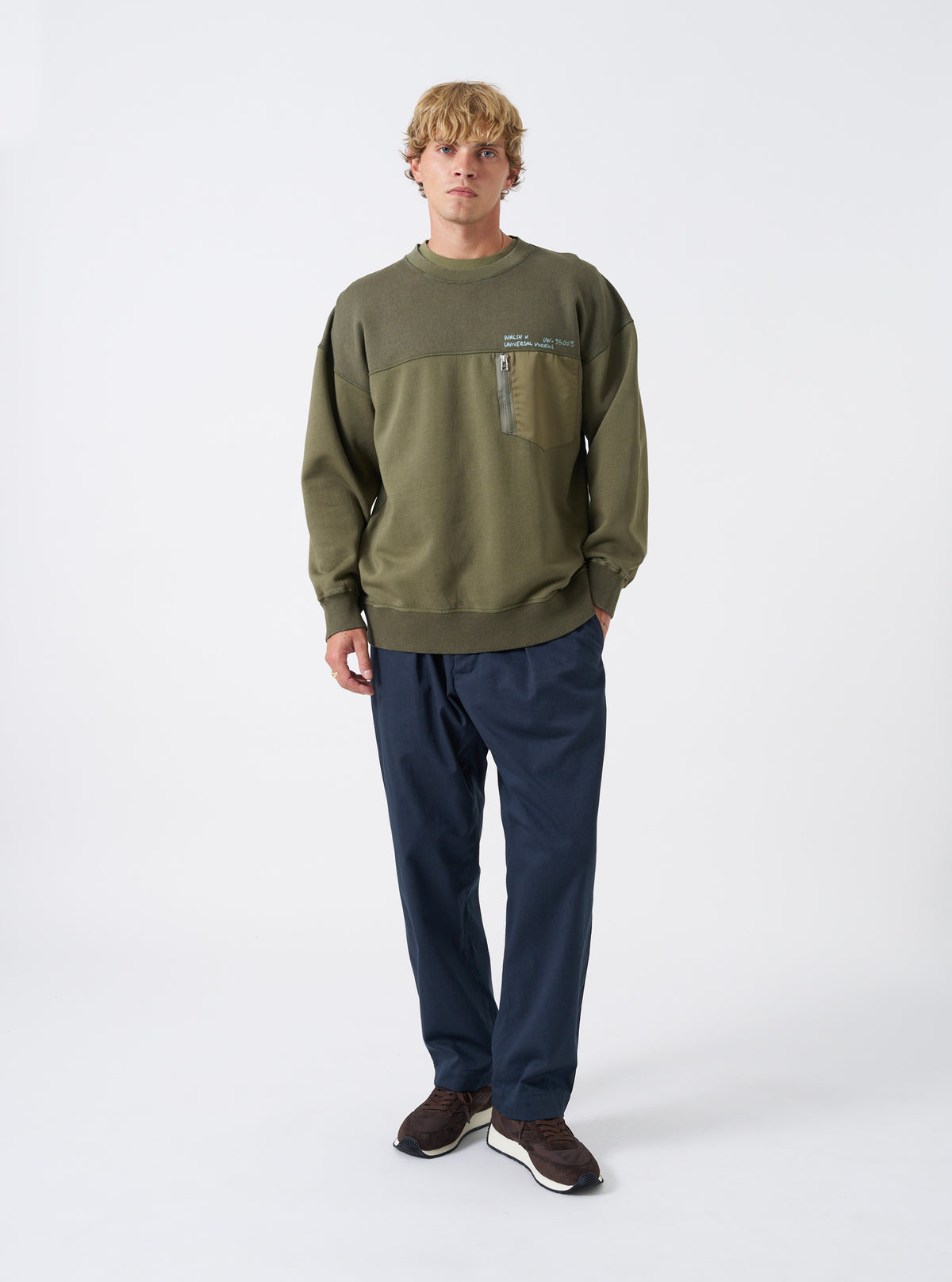 A model wears the Walsh x Universal Works Olive Brushback Sweat Panelled Loose Sweatshirt against a plain studio background. Prominent details are the relaxed fit, olive green tone, crew neckline, and soft brushback cotton texture.