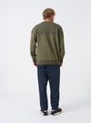 Side perspective of a model styled in the Walsh x Universal Works Olive Brushback Sweat Panelled Loose Sweatshirt in a studio setting. The image shows the loose fit, paneled sleeve detail, ribbed cuffs, and casual olive silhouette.