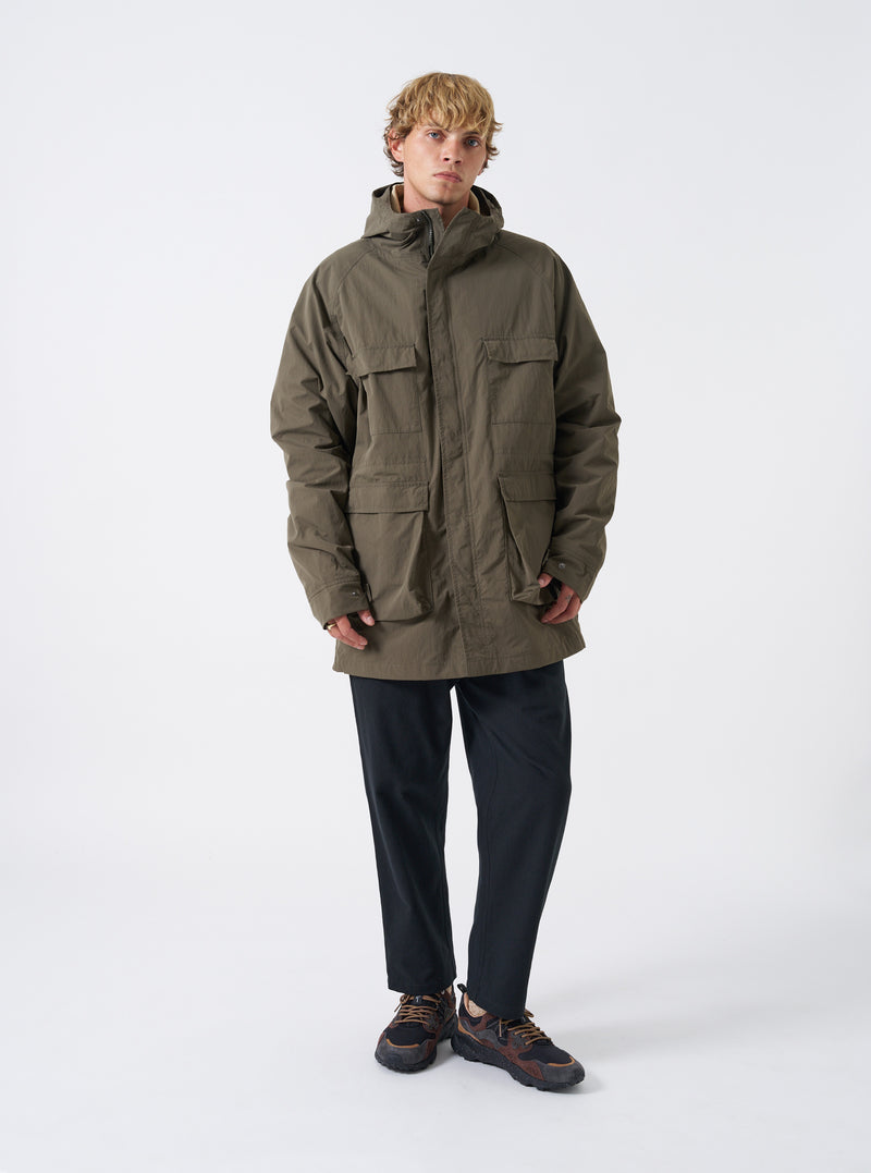 A male model wears the Olive WR Recycled Nylon Peak Parka against a plain studio background. The image showcases the full-length relaxed fit, olive green tone, hooded front design, and functional front cargo pockets.