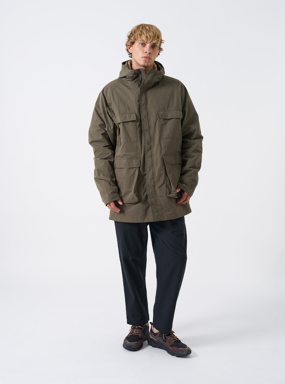 A male model wears the Olive WR Recycled Nylon Peak Parka against a plain studio background. The image showcases the full-length relaxed fit, olive green tone, hooded front design, and functional front cargo pockets.
