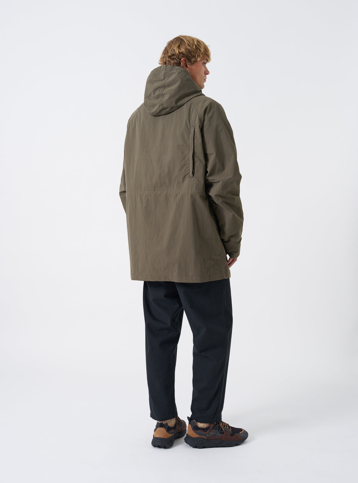 A male model stands against a plain neutral backdrop wearing the Olive WR Recycled Nylon Peak Parka. The photo highlights the back panel silhouette, protective hood, comfortable cut, and mid-length parka hem.