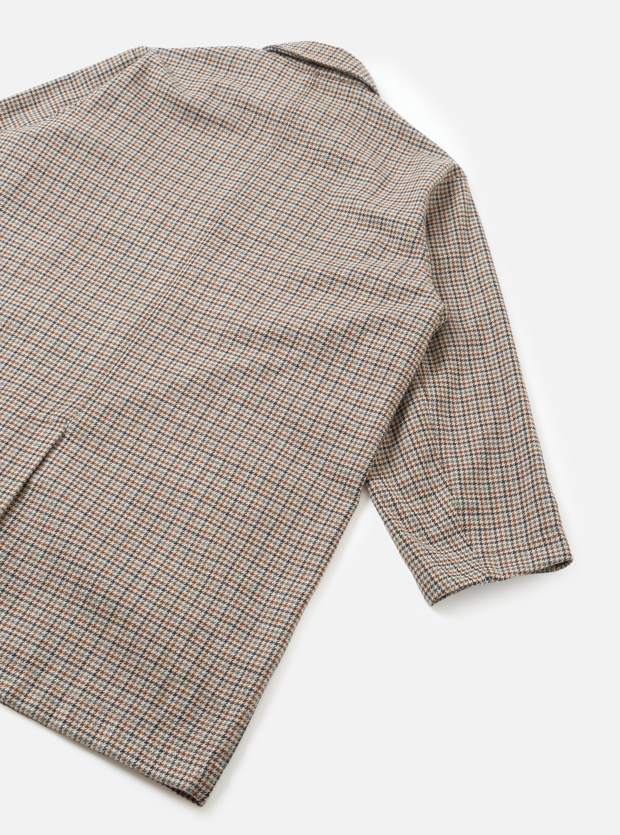 Flat lay detail shot of the taupe Lester Dogtooth Stan Coat on a white studio surface. The image shows the back panel and one long sleeve, highlighting the fine dogtooth weave in taupe, rust, navy and ecru. A notched collar is visible at the top. The cuff is unlined and plain.