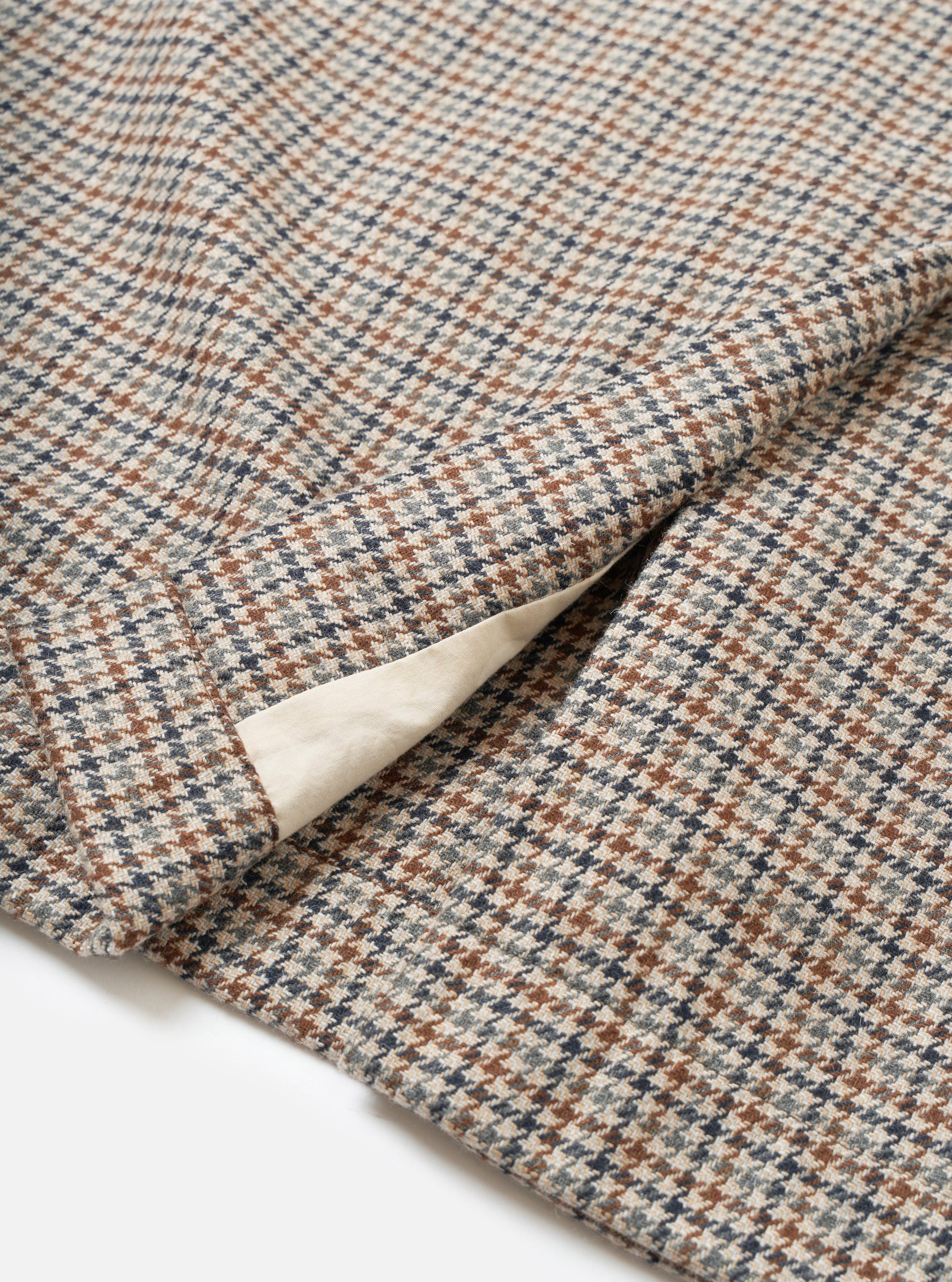 Close-up flat lay of the Lester Dogtooth Stan Coat in taupe, showing the woven dogtooth check fabric in cream, rust brown, and navy. A folded cuff or hem reveals a plain cream lining beneath. Shot on a white studio surface.