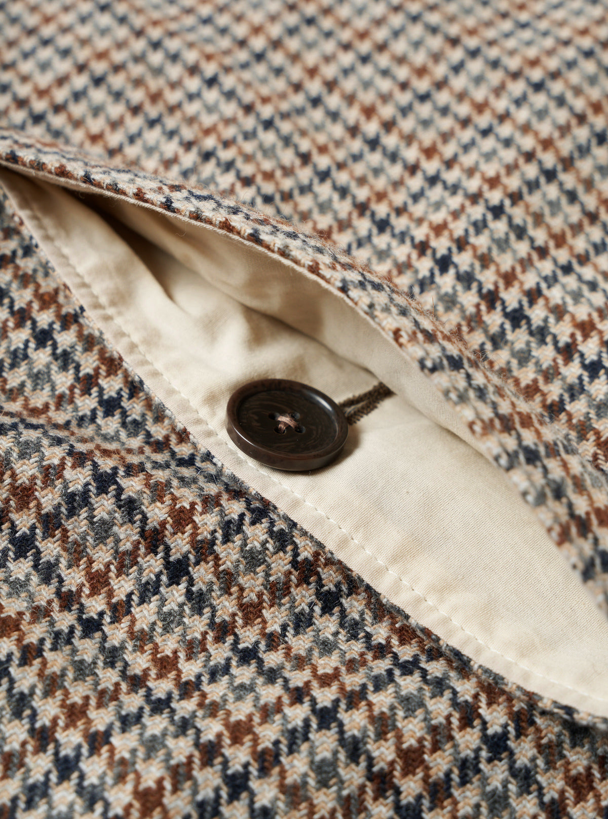 Close-up product view of the Taupe Lester Dogtooth Stan Coat against a plain backdrop. The image emphasizes the textured acrylic-polyester fabric, horn button detail, collar construction, and rich pattern detail.