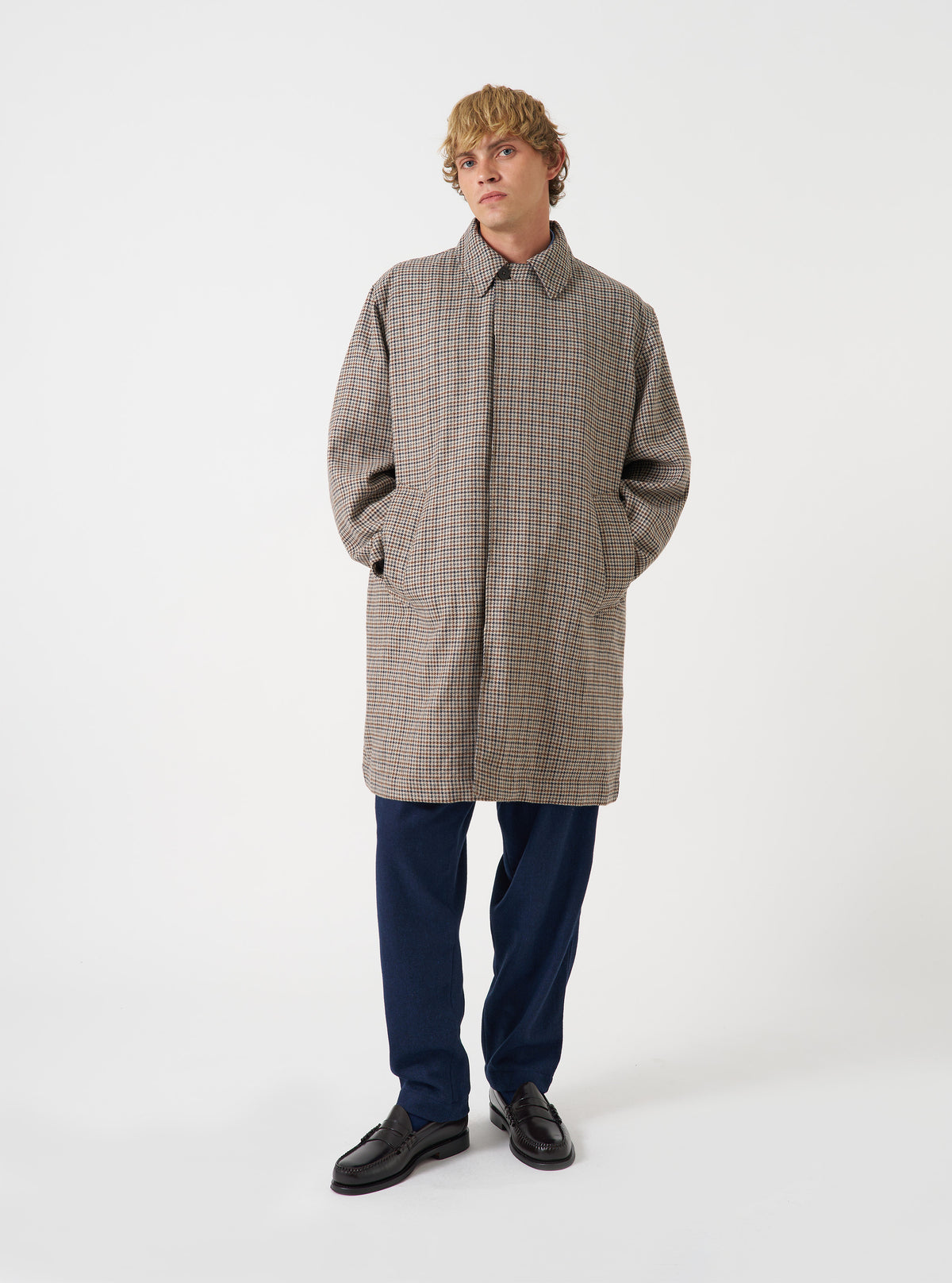 A male model wears the Taupe Lester Dogtooth Stan Coat against a plain neutral background. The photo showcases the long tailored overcoat fit, houndstooth check pattern, collared neckline, and relaxed layering style.