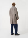 A male model stands in a studio setting with a plain background wearing the Taupe Lester Dogtooth Stan Coat. The image displays the rear profile, gentle drape, brown check weave, and vent detail of the coat.