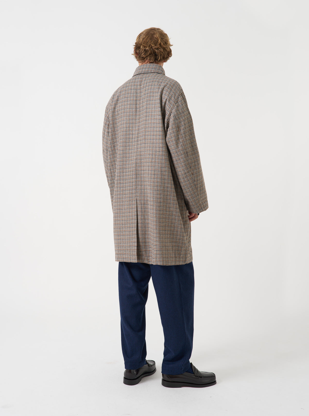 A male model stands in a studio setting with a plain background wearing the Taupe Lester Dogtooth Stan Coat. The image displays the rear profile, gentle drape, brown check weave, and vent detail of the coat.