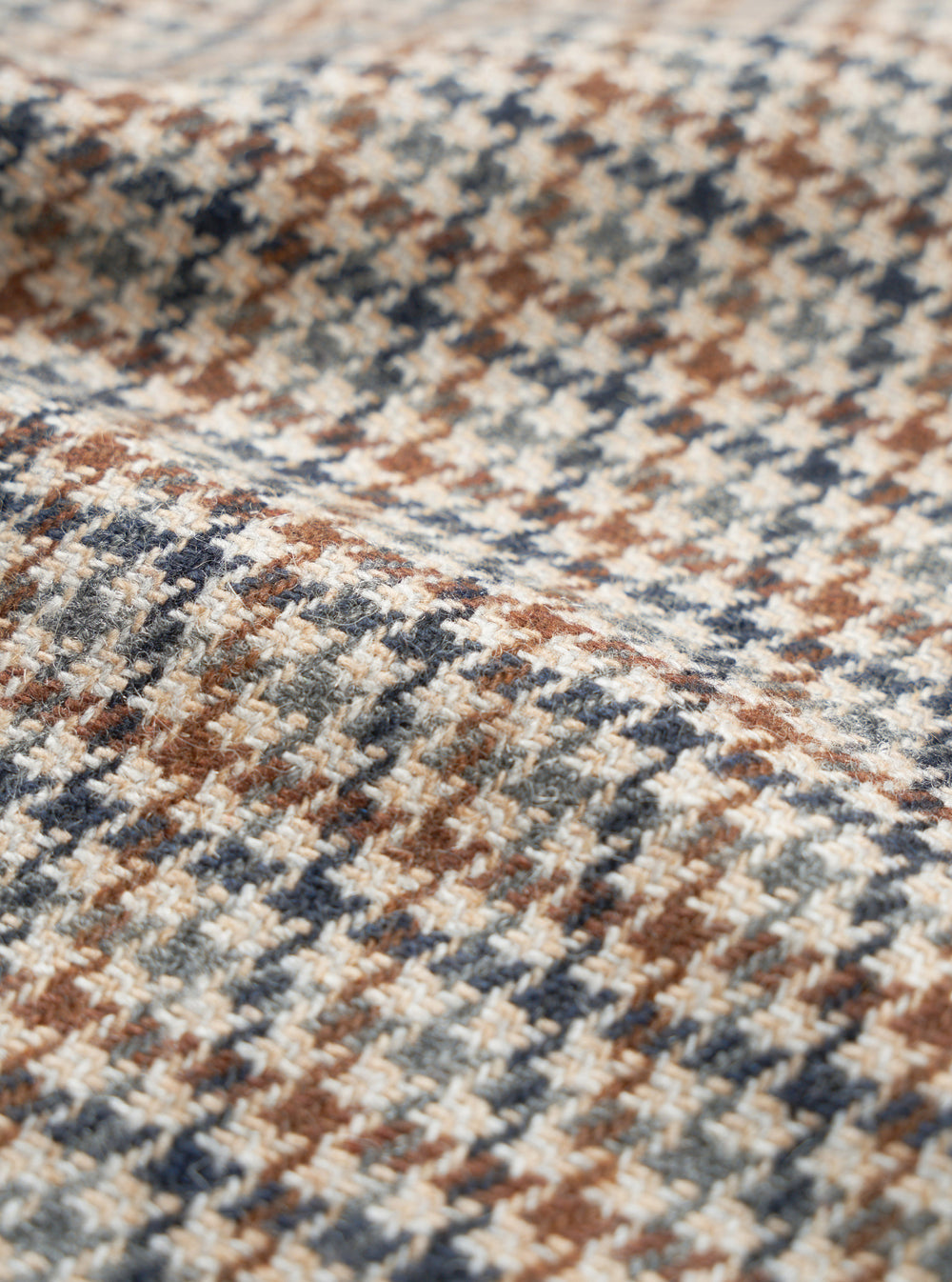Close-up of the taupe Lester Dogtooth Stan Coat fabric, showing a woven dogtooth check pattern in cream, rust brown, navy and grey. The woollen textile has a lightly textured, hairy surface with visible yarn construction. The fabric is gathered into soft folds, highlighting its weight and drape.