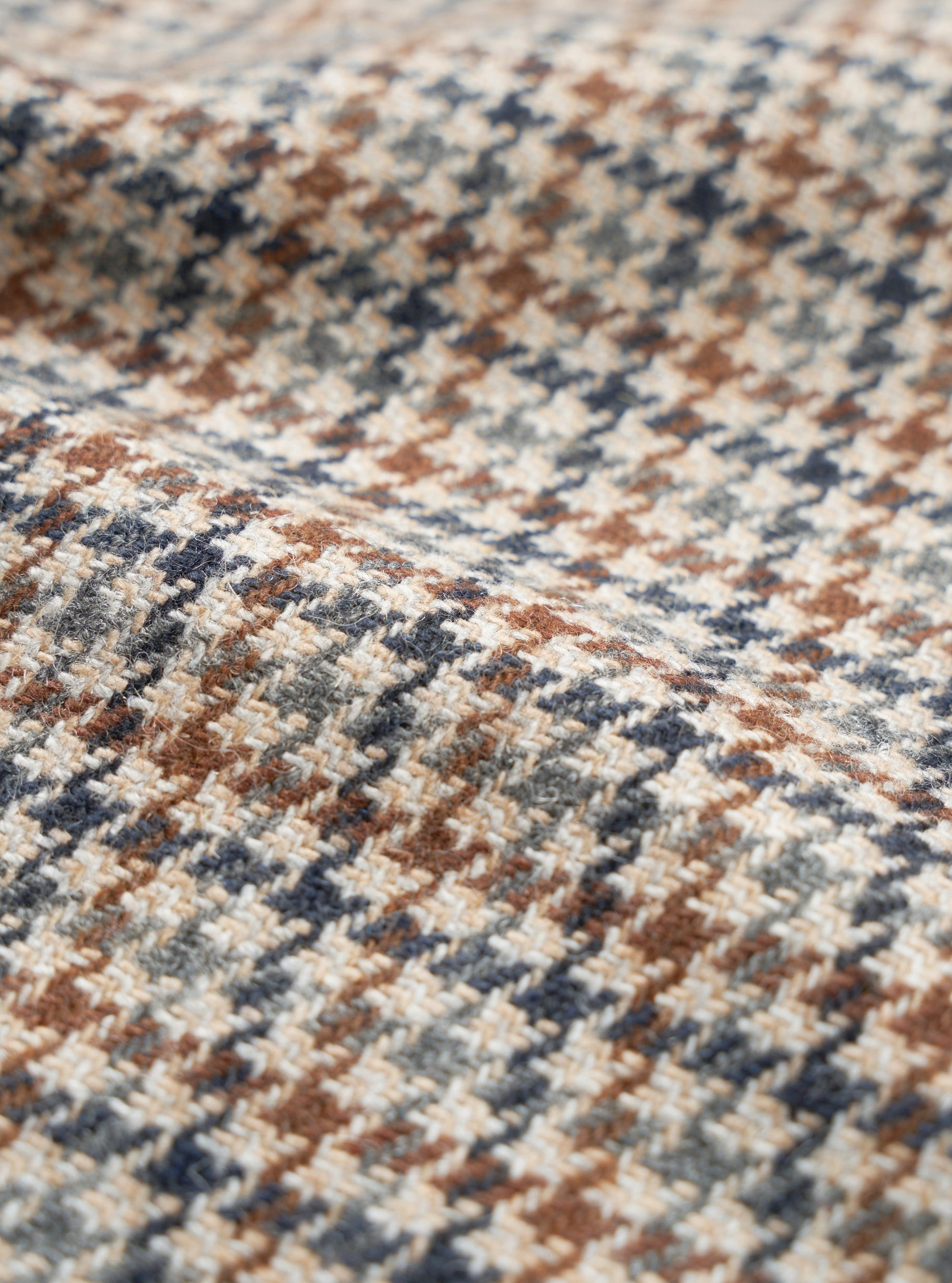 Close-up of the taupe Lester Dogtooth Stan Coat fabric, showing a woven dogtooth check pattern in cream, rust brown, navy and grey. The woollen textile has a lightly textured, hairy surface with visible yarn construction. The fabric is gathered into soft folds, highlighting its weight and drape.