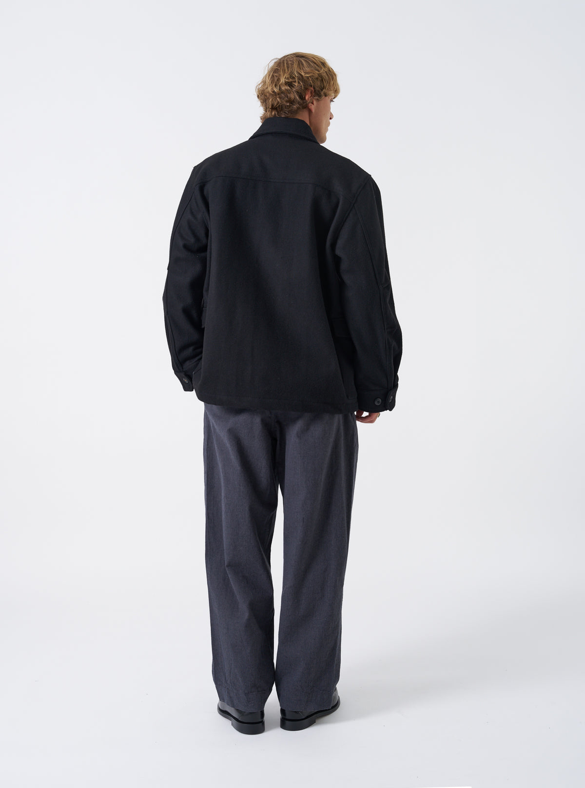A male model stands against a neutral studio backdrop wearing the Black Mowbray Heavy Duty Jacket. The shot displays the back profile, heavy wool-blend texture, comfortable fit, and clean shoulder seams.