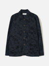 Black wool-mix chore jacket with an abstract inkblot jacquard pattern. Point collar, five dark horn-effect buttons down the centre front, a chest welt pocket and two large patch pockets at the hem. Full-length sleeves with plain cuffs. Universal Works label visible at the neck. Flat lay on a light grey studio backdrop.
