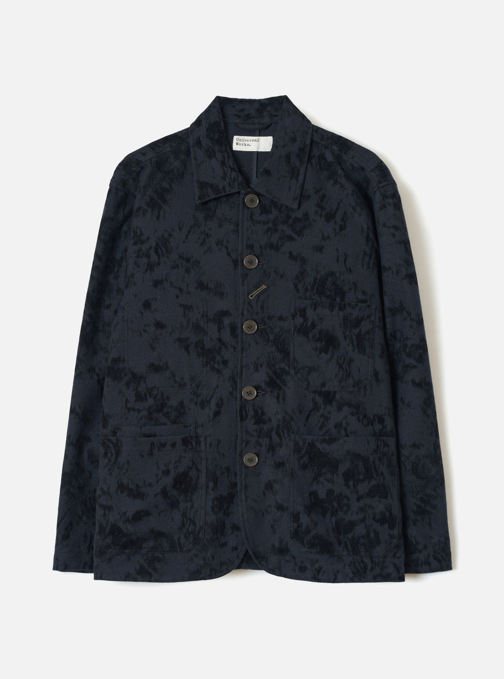 Black wool-mix chore jacket with an abstract inkblot jacquard pattern. Point collar, five dark horn-effect buttons down the centre front, a chest welt pocket and two large patch pockets at the hem. Full-length sleeves with plain cuffs. Universal Works label visible at the neck. Flat lay on a light grey studio backdrop.