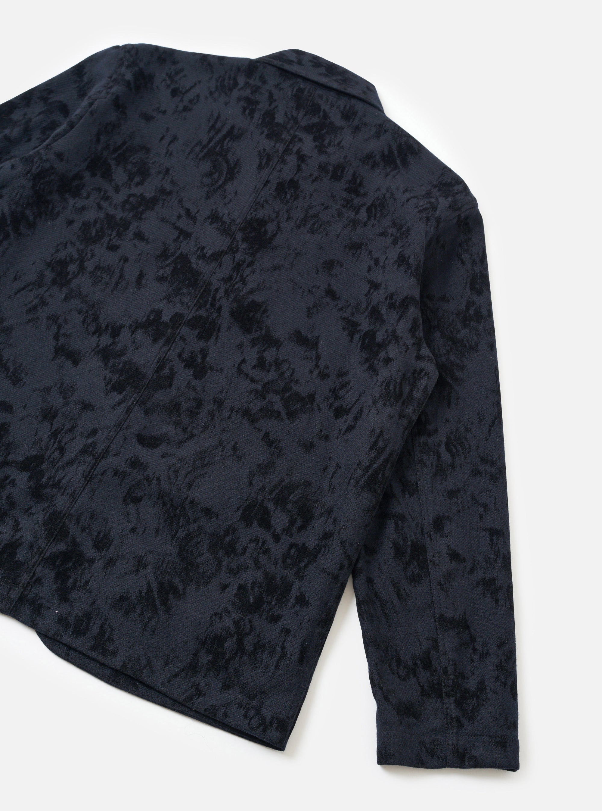 Black wool-mix chore jacket in a bakers cut, laid flat on a white surface, shot from a rear three-quarter angle. The fabric features a tonal abstract floral jacquard pattern in black on navy. A small collar, rear seam, curved hem, and plain long sleeve cuff are visible.