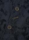 Close-up of the front button placket on a navy wool mix chore jacket with an all-over abstract jacquard pattern in black. Two large four-hole horn-effect buttons are visible, with a gold chain stitch buttonhole between them. The fabric has a textured, brushed surface with diagonal tonal markings.
