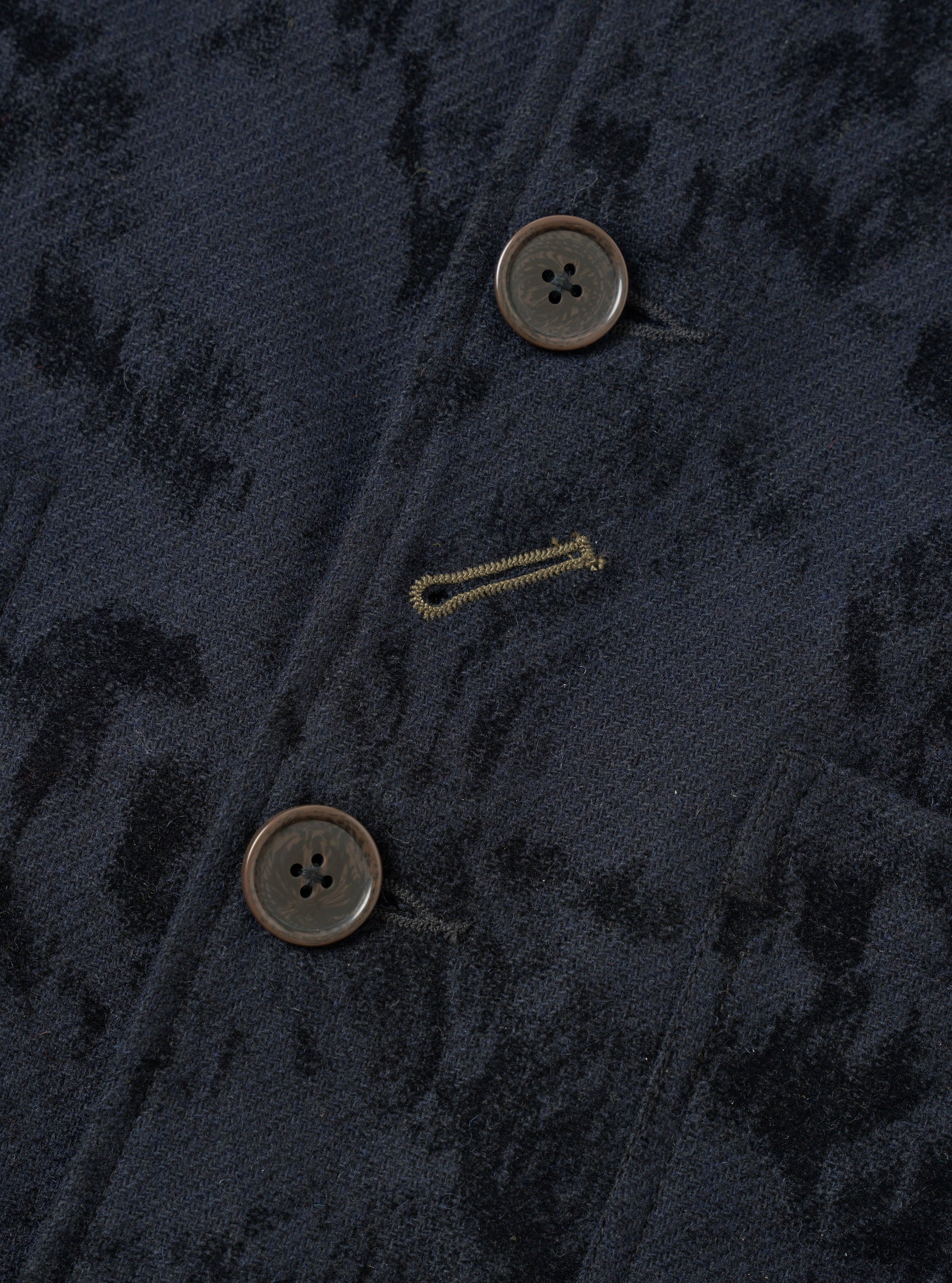 Close-up of the front button placket on a navy wool mix chore jacket with an all-over abstract jacquard pattern in black. Two large four-hole horn-effect buttons are visible, with a gold chain stitch buttonhole between them. The fabric has a textured, brushed surface with diagonal tonal markings.