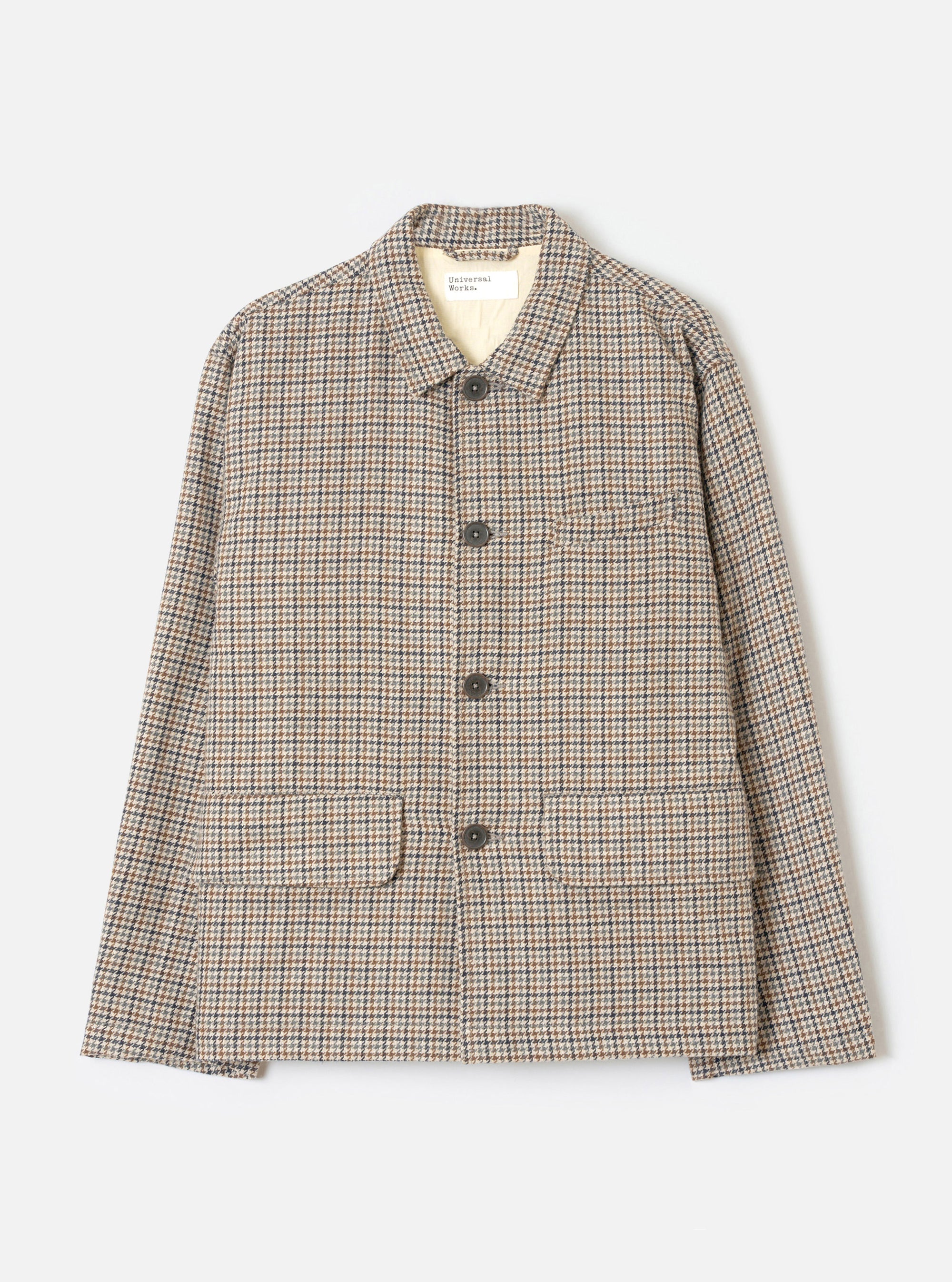 Taupe dogtooth Franck jacket laid flat on a light grey background. The wool-mix fabric features a multicolour dogtooth pattern in taupe, navy and rust. Point collar with a Universal Works woven label at the neck. Three dark horn buttons down the centre front. One chest welt pocket and two large patch pockets at the hem.