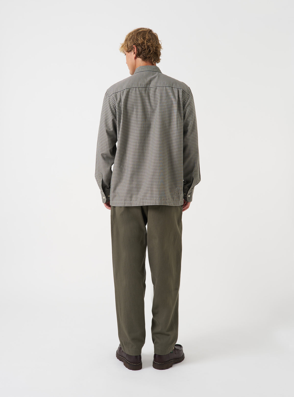 A rear view of a model wearing the Universal Works Olive Brushed Moleskin Pleated Track Pant set against a plain backdrop. The image highlights the olive green brushed fabric, buttoned back pockets, and narrow hem of the trousers.