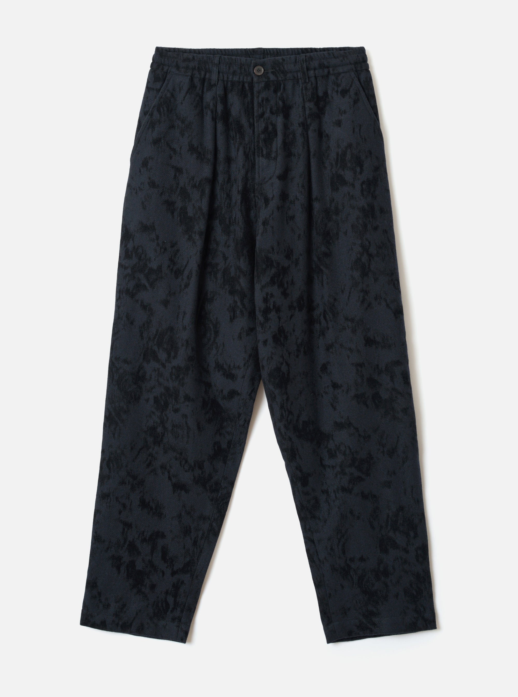 Black wool-mix track pants with an all-over abstract blurred print in dark navy and black tones. Features an elasticated waistband with a single centre button, twin pleats at the front, side slash pockets, and a relaxed tapered leg to an unfinished hem. Flat lay on a light grey studio backdrop.