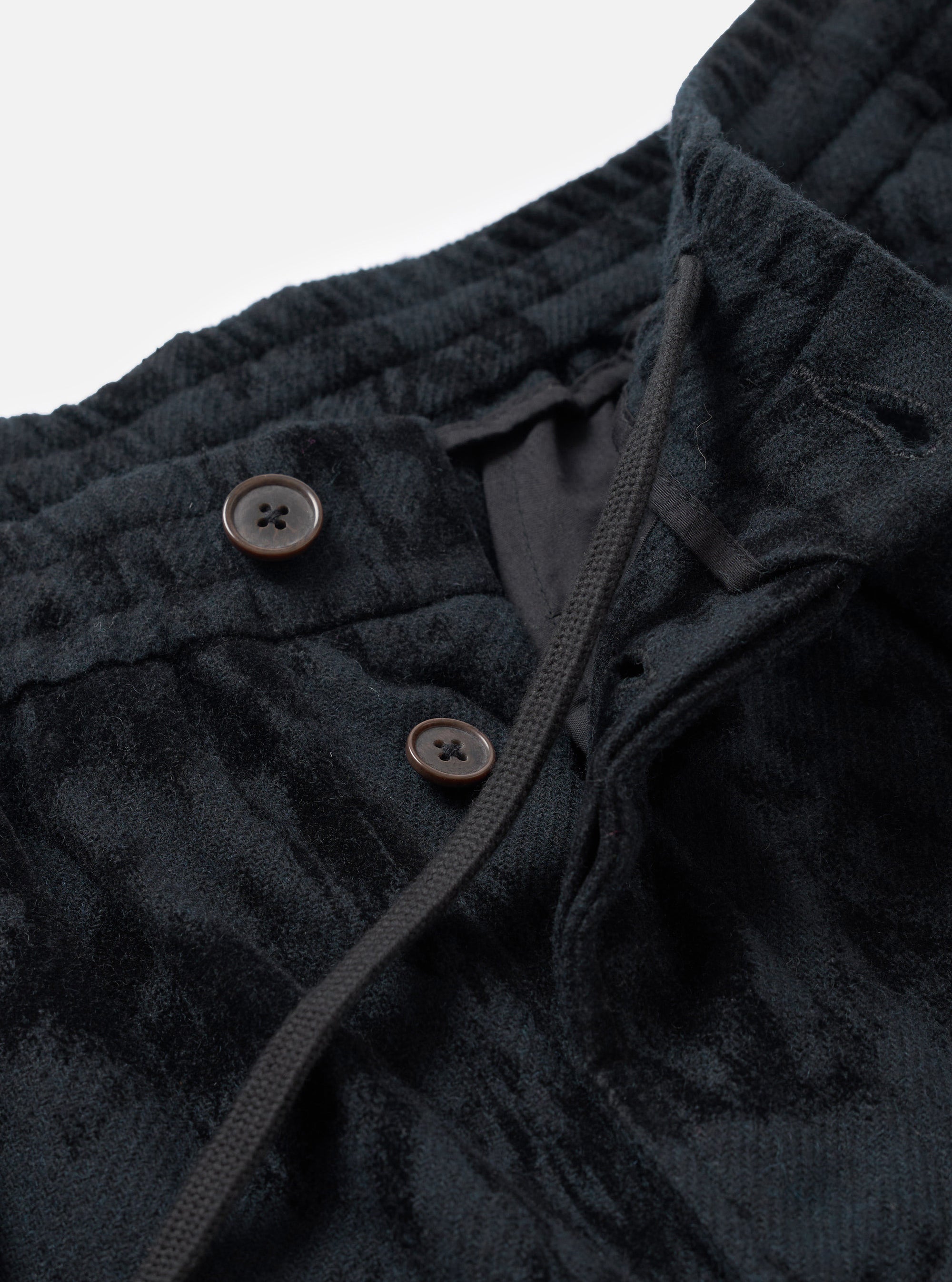 Close-up detail of the black wool mix pleated track pant's waistband and fly area. Two dark brown four-hole buttons fasten the placket. A woven drawcord threads through the ribbed elasticated waistband. The textured fabric shows a tonal check pattern. Shot against a plain light grey studio backdrop.