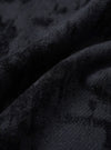 Close-up detail of the black wool mix fabric used in the pleated track pant. The surface has a dense, lightly boiled or brushed texture with fine fibres visible throughout. Deep folds in the fabric reveal soft drape and a matte finish with subtle tonal variation across the dark charcoal-black cloth.