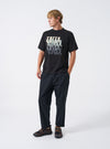 A model wearing the Universal Works Black Brushed Moleskin Hi Water Trouser, shown from the front against a plain background. The black cotton pants feature a cropped ankle length, elasticated drawstring waistband, and slim tapered fit.