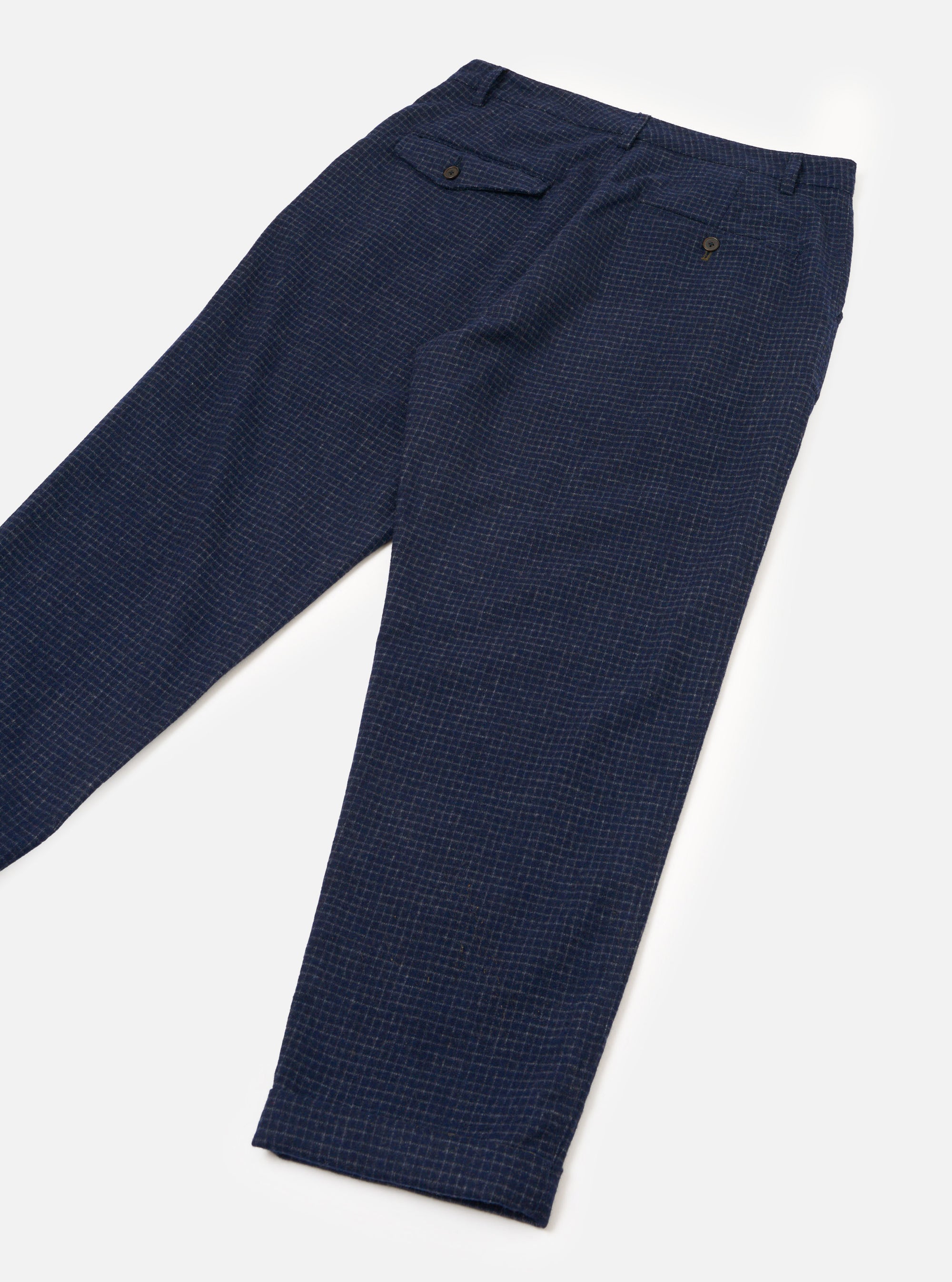Navy check wool mix chinos laid flat on a white surface, angled diagonally. Fine light-coloured grid check on a navy ground with a textured weave. Waistband with belt loops visible, a buttoned flap pocket at the left hip, a rear welt pocket with a dark button, and an unfinished hem at the ankle.