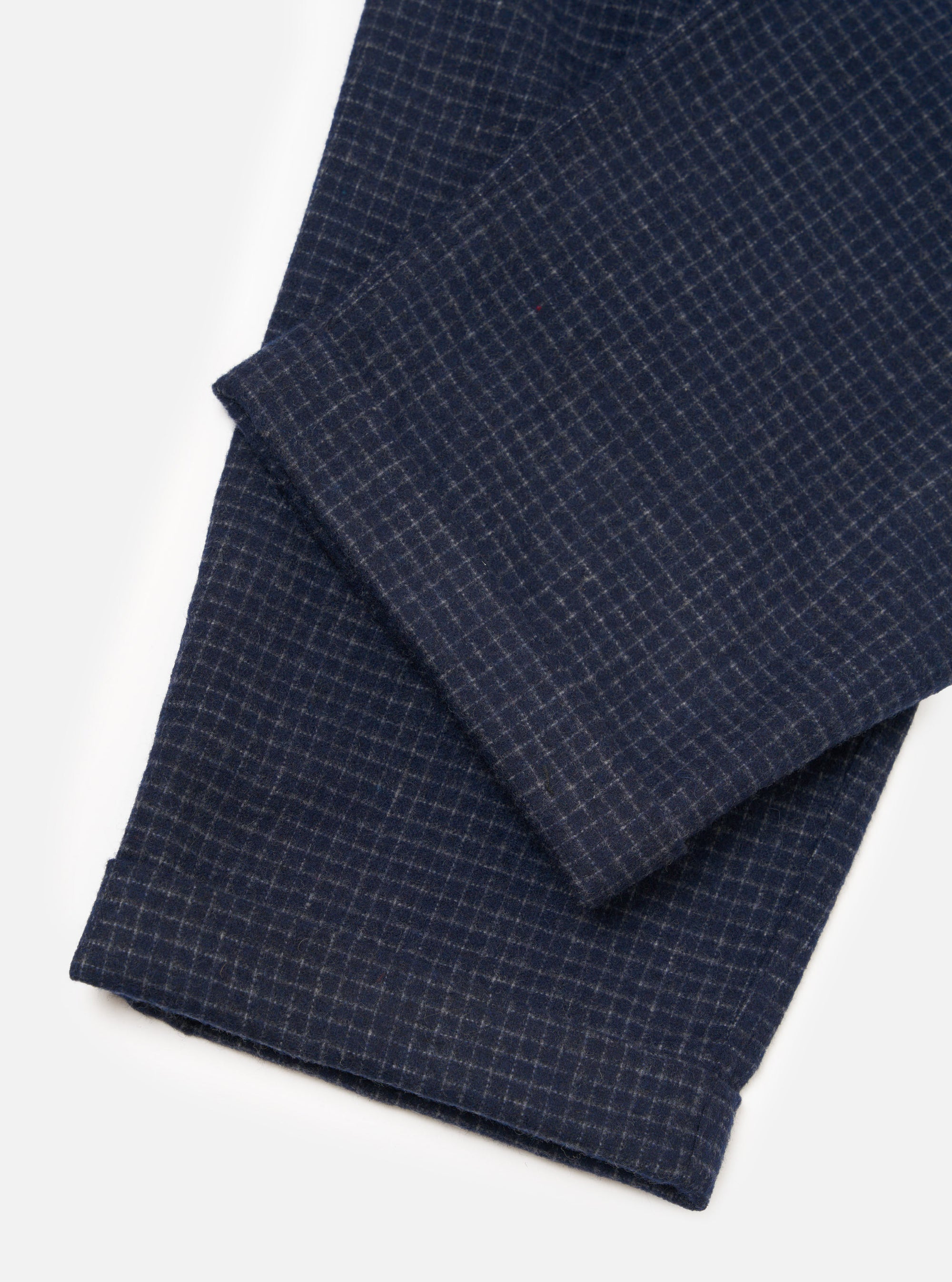 Close-up flat lay of the navy check Bobby Wool Mix Super Chino on a white studio surface, showing the lower leg and turned-up cuff. The fabric features a fine grey grid check on a navy ground with a lightly textured, wool-mix weave. The cuff fold and finished hem are clearly visible.