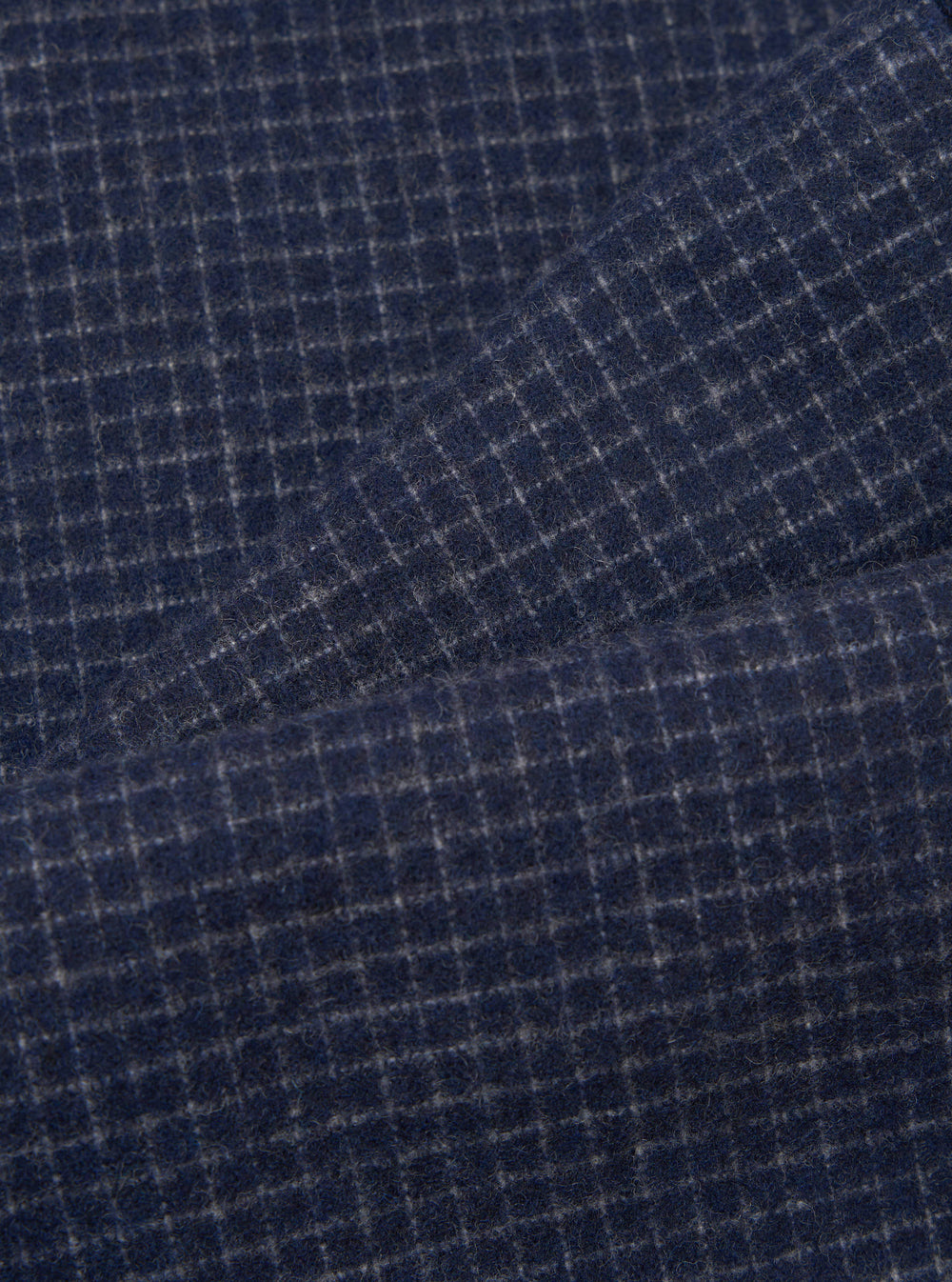 Close-up fabric shot of the navy check wool mix super chino, showing a fine grey windowpane check pattern on a deep navy ground. The brushed, slightly hairy texture of the wool mix fabric is clearly visible. A diagonal fold runs across the upper portion of the frame, revealing the fabric's drape.