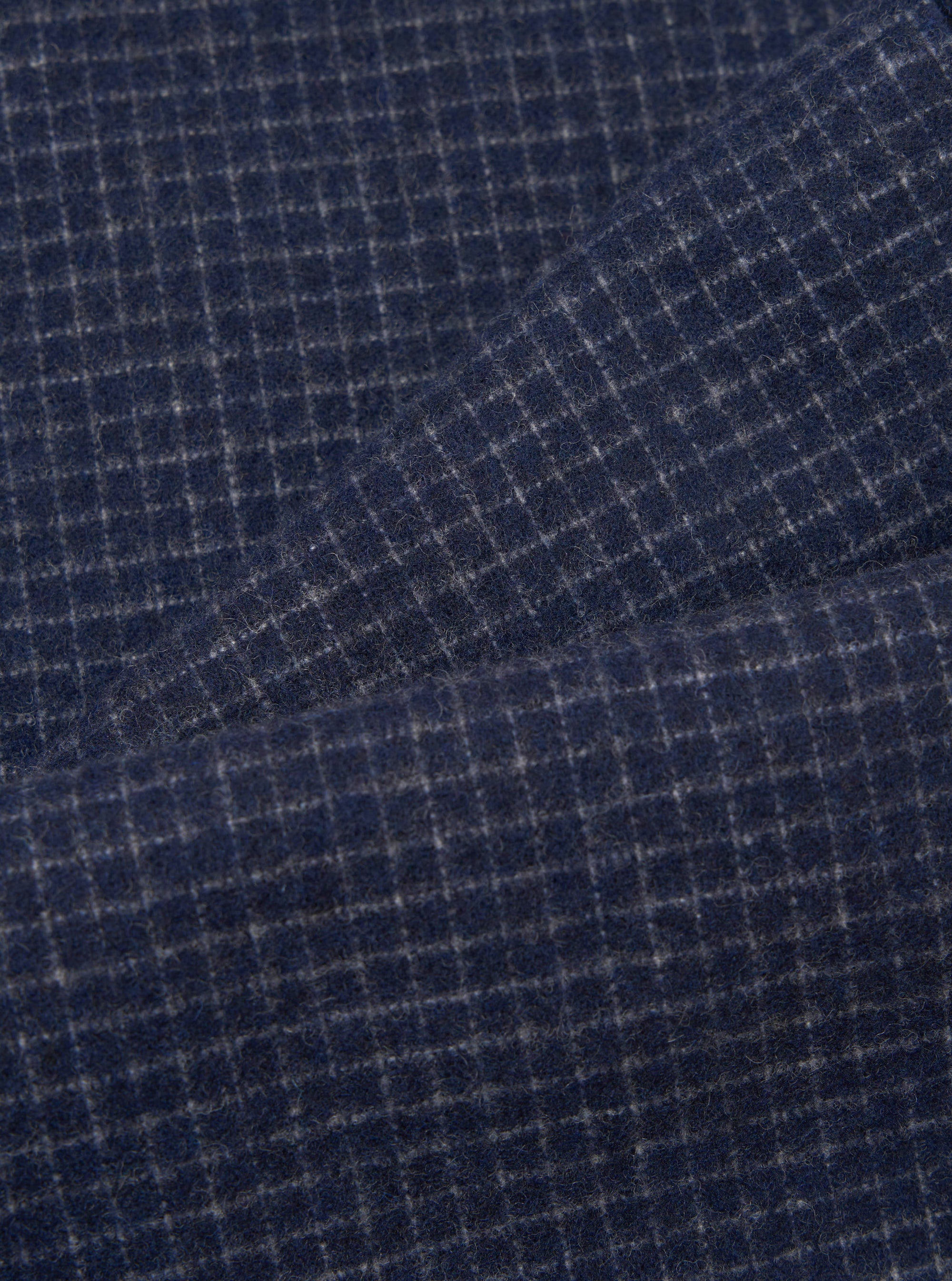 Close-up fabric shot of the navy check wool mix super chino, showing a fine grey windowpane check pattern on a deep navy ground. The brushed, slightly hairy texture of the wool mix fabric is clearly visible. A diagonal fold runs across the upper portion of the frame, revealing the fabric's drape.