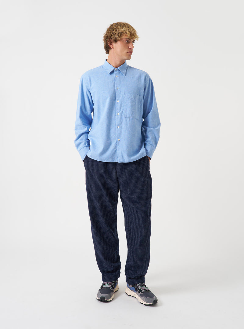 A model wearing the Universal Works Navy Check Bobby Wool Mix Super Chino, viewed from the front against a plain backdrop. The trousers showcase a navy and ecru windowpane check pattern on brushed wool-blend fabric with cuffed hems.