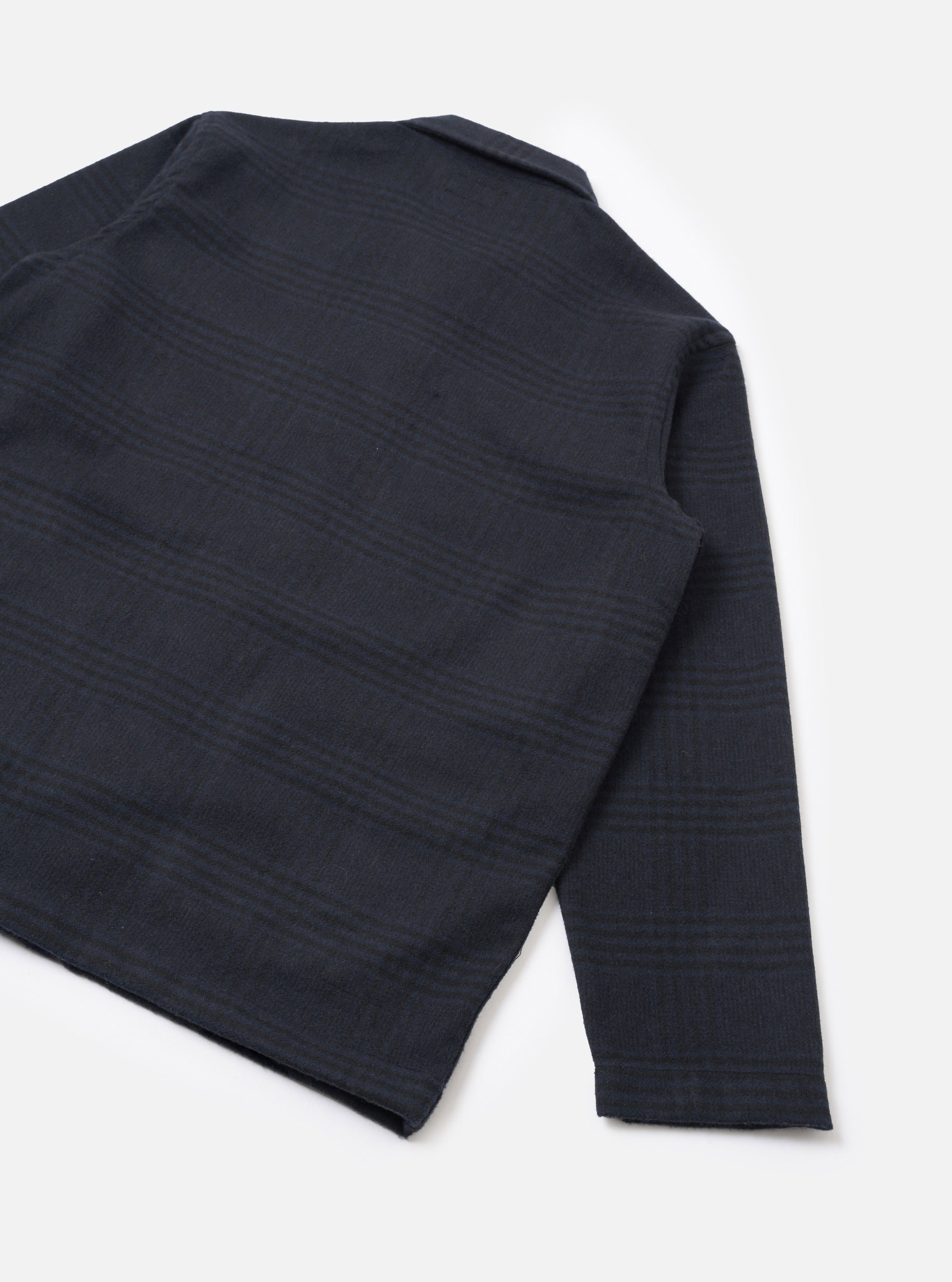 Flat lay of the navy wool mix bakers overshirt on a white studio surface, showing the back and one sleeve at an angle. The fabric features a tonal check pattern woven into the dark navy ground. The straight hem and unadorned cuff are visible, with a small collar visible at the top.