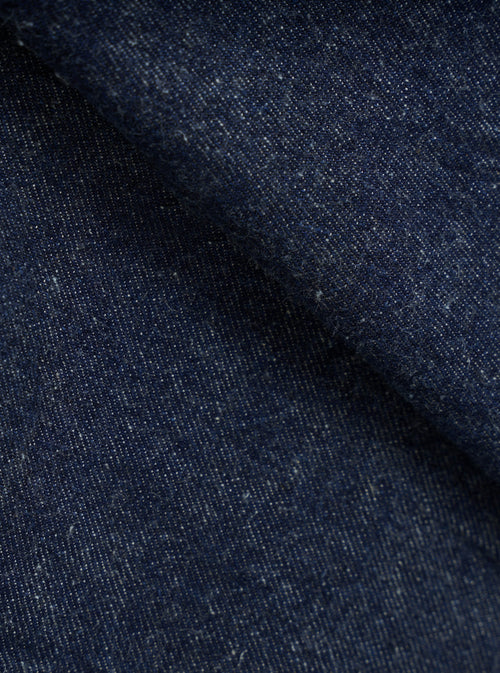 Close-up of indigo denim fabric showing a textured twill weave with flecks of white yarn throughout. The deep navy blue cloth is folded, creating soft ridges that highlight the fabric's weight and slub texture. No garment construction details are visible.