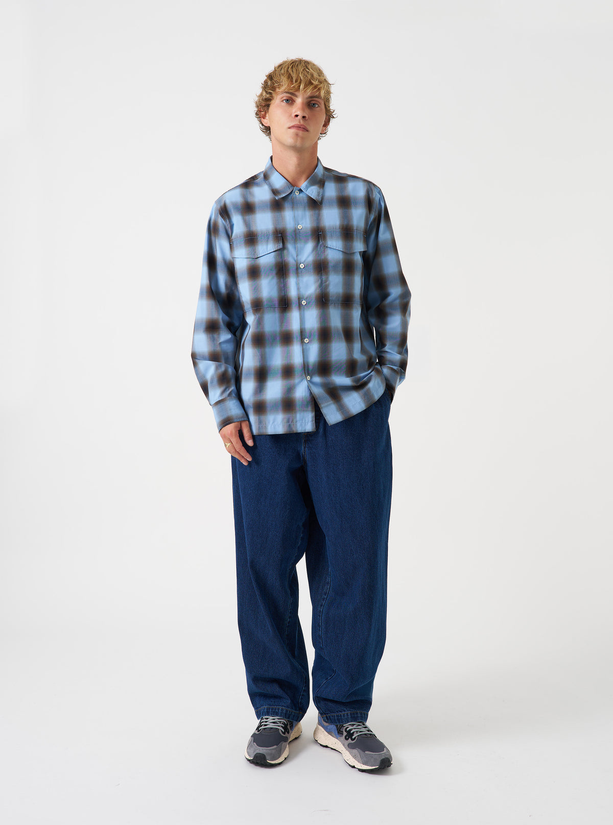 A model wearing the Universal Works Blue Check Charlie Check Attendant Shirt, viewed from front. The long-sleeve shirt features a blue check pattern, flat collar, central button closure, and two large chest flap pockets against a plain backdrop.