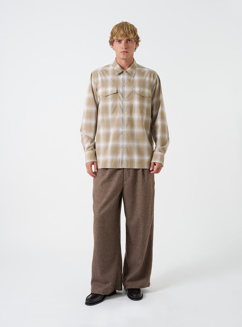 A model wearing the Universal Works Sand Check Charlie Check Attendant Shirt, captured from the front. The shirt displays a sand-toned check pattern, flat collar, button-down placket, and prominent chest flap pockets against a plain backdrop.
