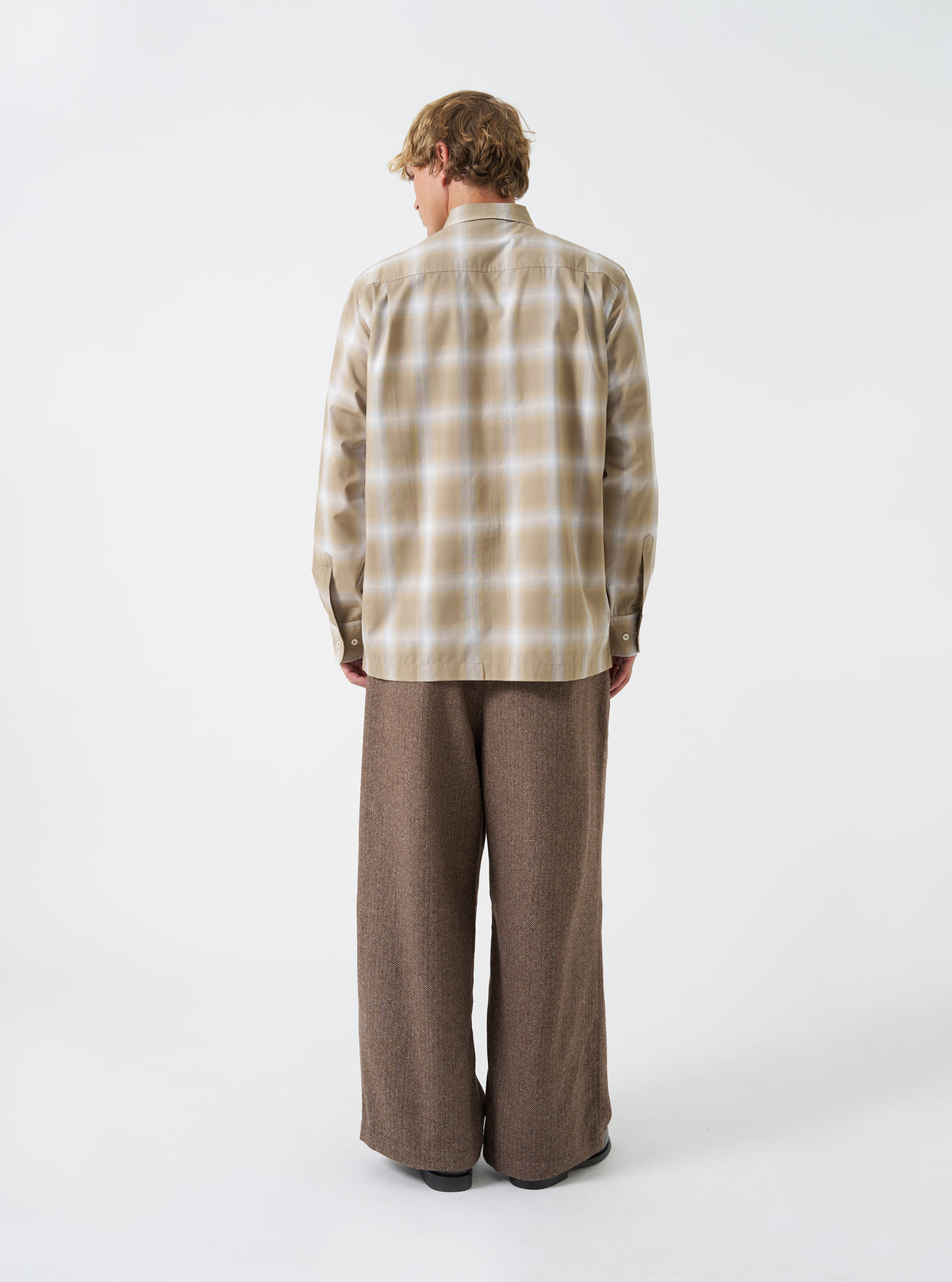 A back view of a model wearing the Universal Works Sand Check Charlie Check Attendant Shirt. The image shows the rear cut of the sand check patterned long-sleeve shirt, highlighting the clean stitching and relaxed hem set against a plain backdrop.