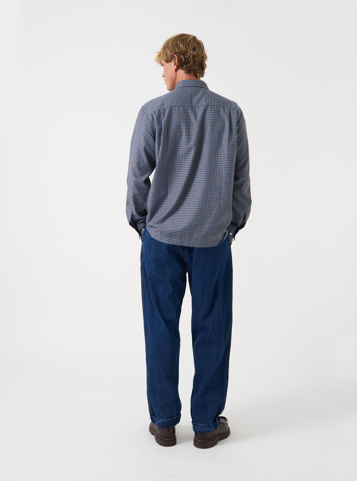 A back view of a model wearing the Universal Works Blue/Brown Parker Check Attendant Shirt. The photo highlights the brown and blue check pattern across the back, straight hemline, and casual silhouette set against a plain backdrop.