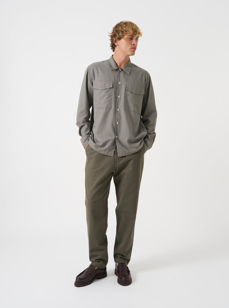 A model wearing the Universal Works Sand/Olive Parker Check Attendant Shirt, viewed from the front. The long-sleeve shirt exhibits a sand and olive check design, flat collar, button closure, and chest patch flap pockets against a plain background.
