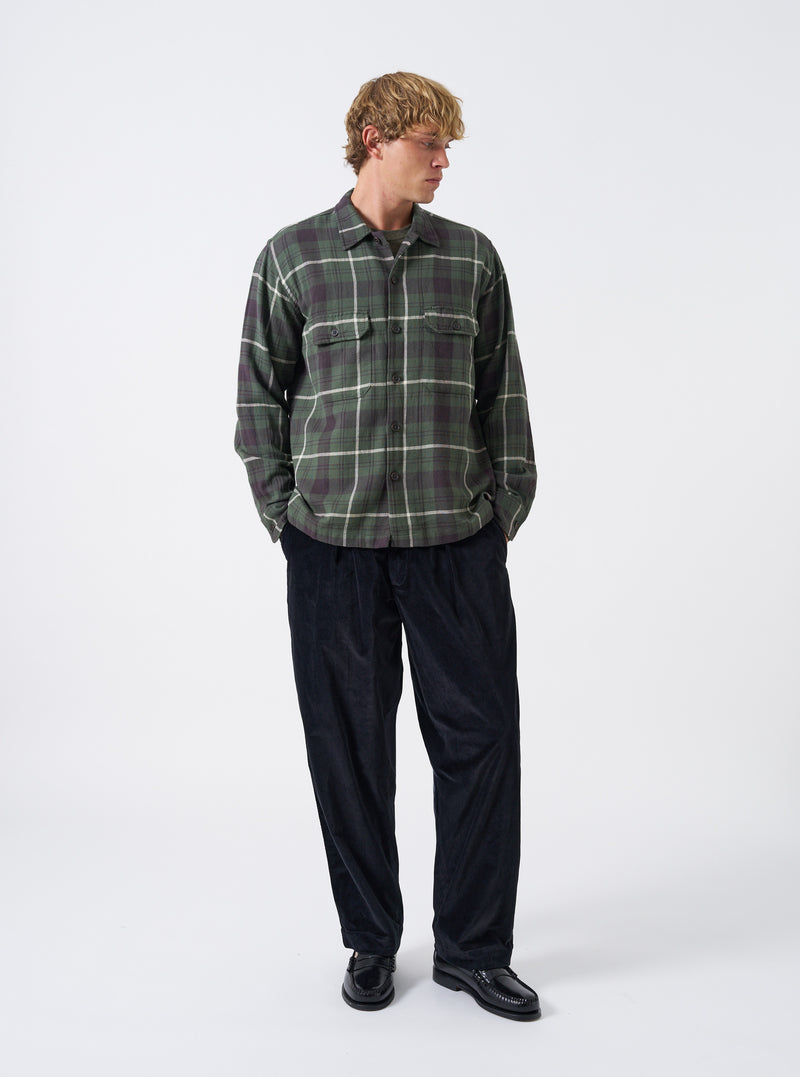 A model wearing the Universal Works Olive Washed Check L/S Utility Shirt, captured from the front. The long-sleeve shirt showcases an olive green check pattern, flat collar, central buttoning, and dual flap chest pockets against a plain backdrop.