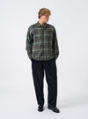 A model wearing the Universal Works Olive Washed Check L/S Utility Shirt, captured from the front. The long-sleeve shirt showcases an olive green check pattern, flat collar, central buttoning, and dual flap chest pockets against a plain backdrop.