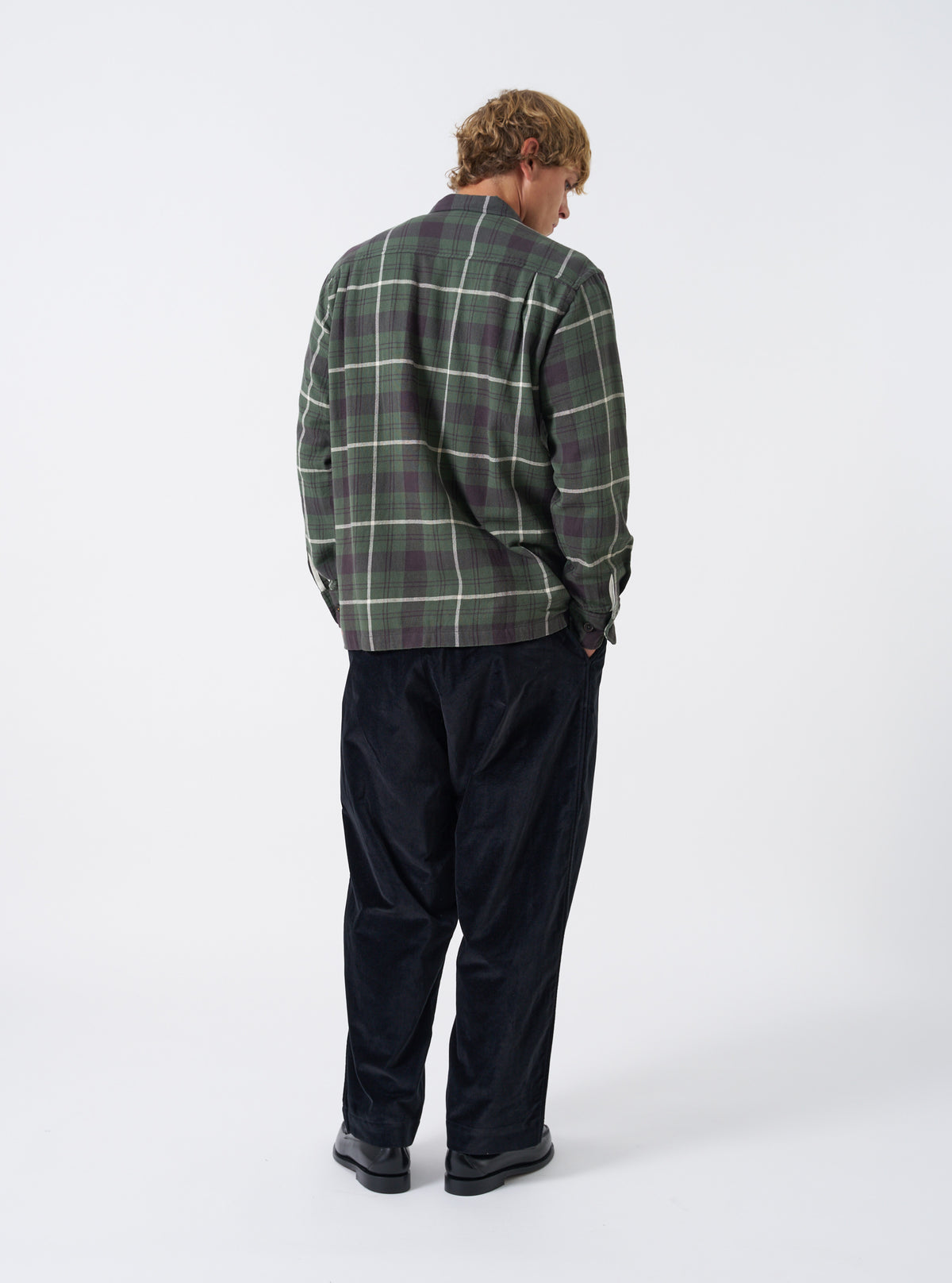 A back view of a model wearing the Universal Works Olive Washed Check L/S Utility Shirt. The photograph shows the rear panel with its olive washed check pattern, easy-fitting shoulders, and straight hemline set against a plain background.